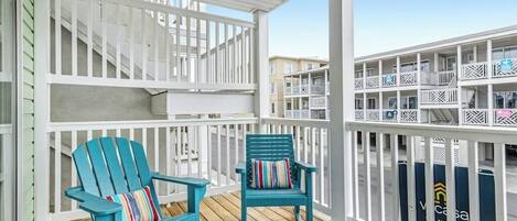 A covered outdoor area with two blue Adirondack chairs, white railings, and a view of a multi-story building.