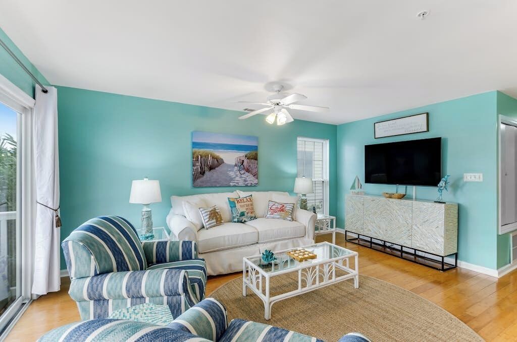 A living room with a teal wall, a white ceiling fan, a large TV, and a sofa with striped cushions.