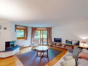 A modern living room with a wooden floor, a round table, a sofa, a television, and a sliding glass door leading to an outdoor area. - Apartment Trais Tschiervs by Interhome (St. Moritz)