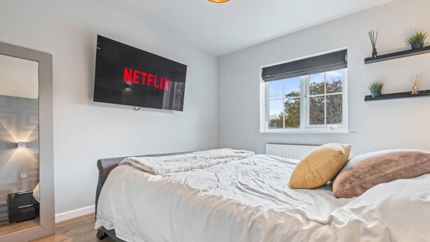 Ett sovrum med en säng, en väggmonterad TV som visar ”NETFLIX”, ett fönster med utsikt och en hylla med diverse föremål.