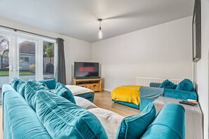Ein modernes Wohnzimmer mit einer Sofagarnitur, einem Fernseher auf einem Holzständer und Blick durch eine Glastür.