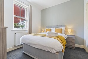 A bedroom with a large bed, a bedside table with a lamp, a window with curtains, and a radiator. - Central Derby Stay | Comfort for 4 | Wi-Fi (Derby)