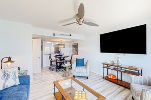 Un salon doté d’un ventilateur de plafond, d’un téléviseur à écran plat fixé au mur, d’un canapé avec un coussin, d’une table basse et d’un coin repas avec des chaises et une table.