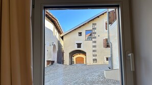 Une vue à travers une fenêtre sur une rue étroite pavée de pierres, avec une porte en bois et des bâtiments à l’architecture traditionnelle.