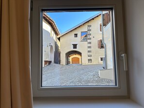 Blick durch ein Fenster auf eine enge Straße mit Kopfsteinpflaster, einer Holztür und Häusern mit traditioneller Architektur.
