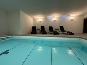 Ein Swimmingpool mit Liegestühlen und einem kleinen Tisch, umgeben von schlichten Wänden und eingelassener Beleuchtung.