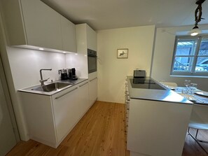 Eine kleine Küche mit weißen Schränken, einem Edelstahl-Spülbecken, einer Kaffeemaschine und einem Fenster mit Blick auf einen Parkplatz.