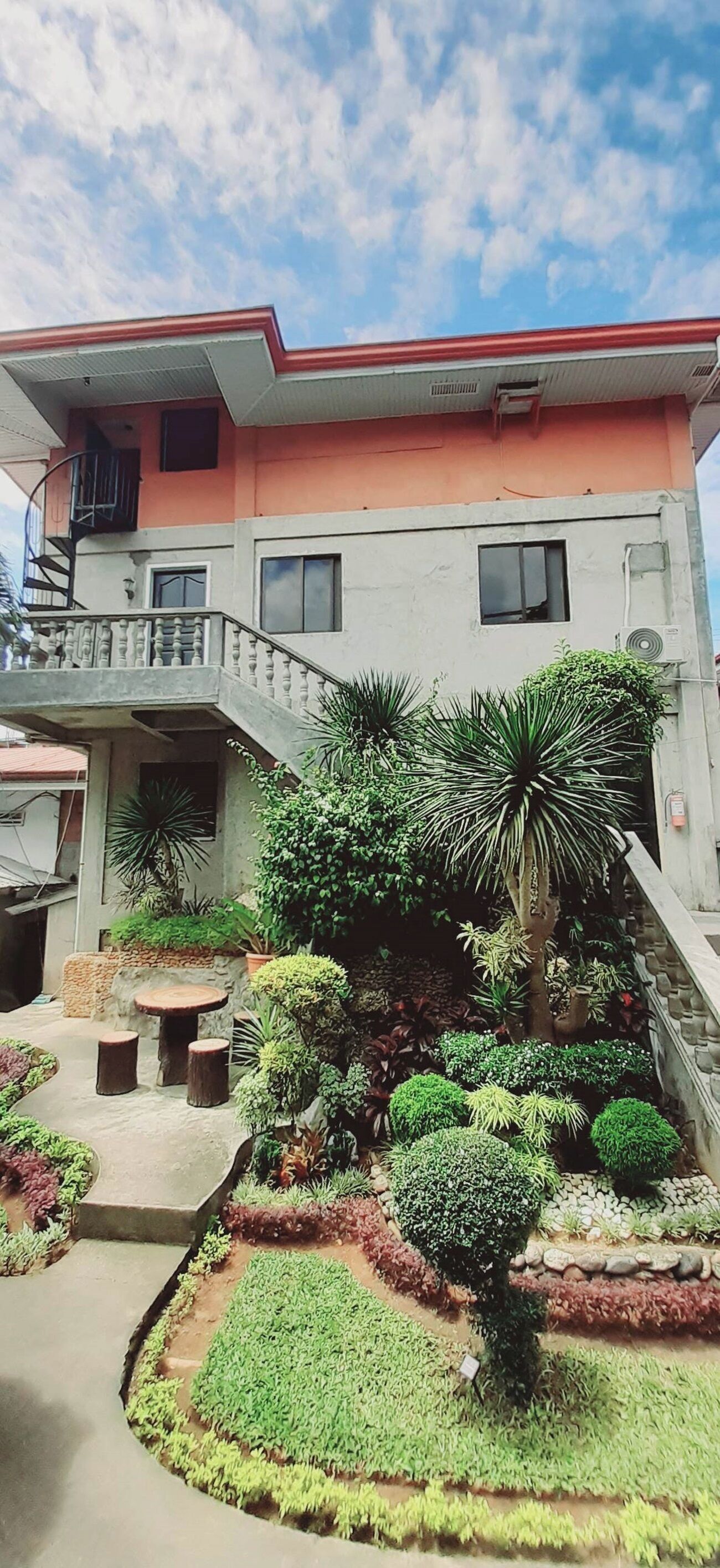 A two-story building with a balcony, a small garden with various plants, and a staircase leading to the upper floor.