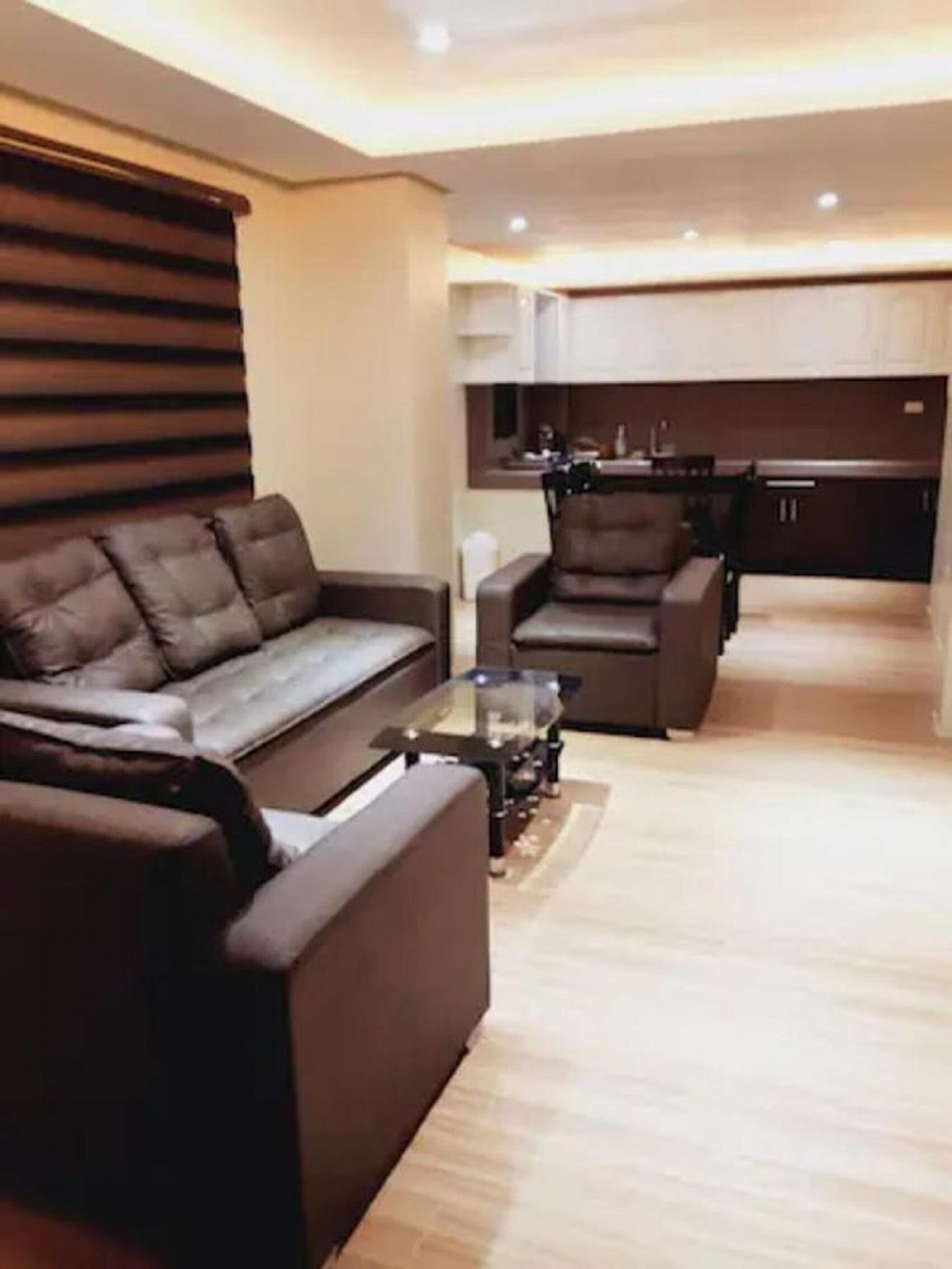 Una sala moderna con un sofá y sillones, una mesa de centro de vidrio, y un área de cocina con armarios y encimera.