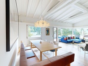 Een ruime woonkamer met een houten eettafel, banken, een blauwe bank en een groot raam met uitzicht op het omliggende landschap.
