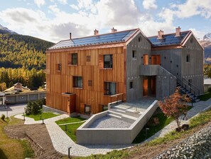 A modern two-story building with a wooden facade and solar panels on the roof, surrounded by a landscaped garden and a mountainous backdrop. - Apartment Julier Nouva by Interhome (Pontresina)