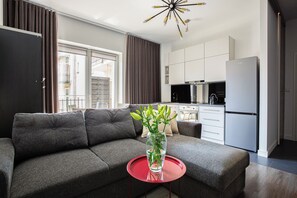 Un salon moderne avec un canapé gris, une table d’appoint rouge ornée d’un vase, et un coin cuisine avec des meubles blancs et un réfrigérateur gris.