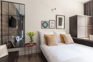 Ein modernes Hotelzimmer mit einem großen Spiegel, einem Bett mit weißen und beigen Kissen, einem kleinen Nachttisch mit Blumen in einer Vase und einem gerahmten Bild an der Wand.