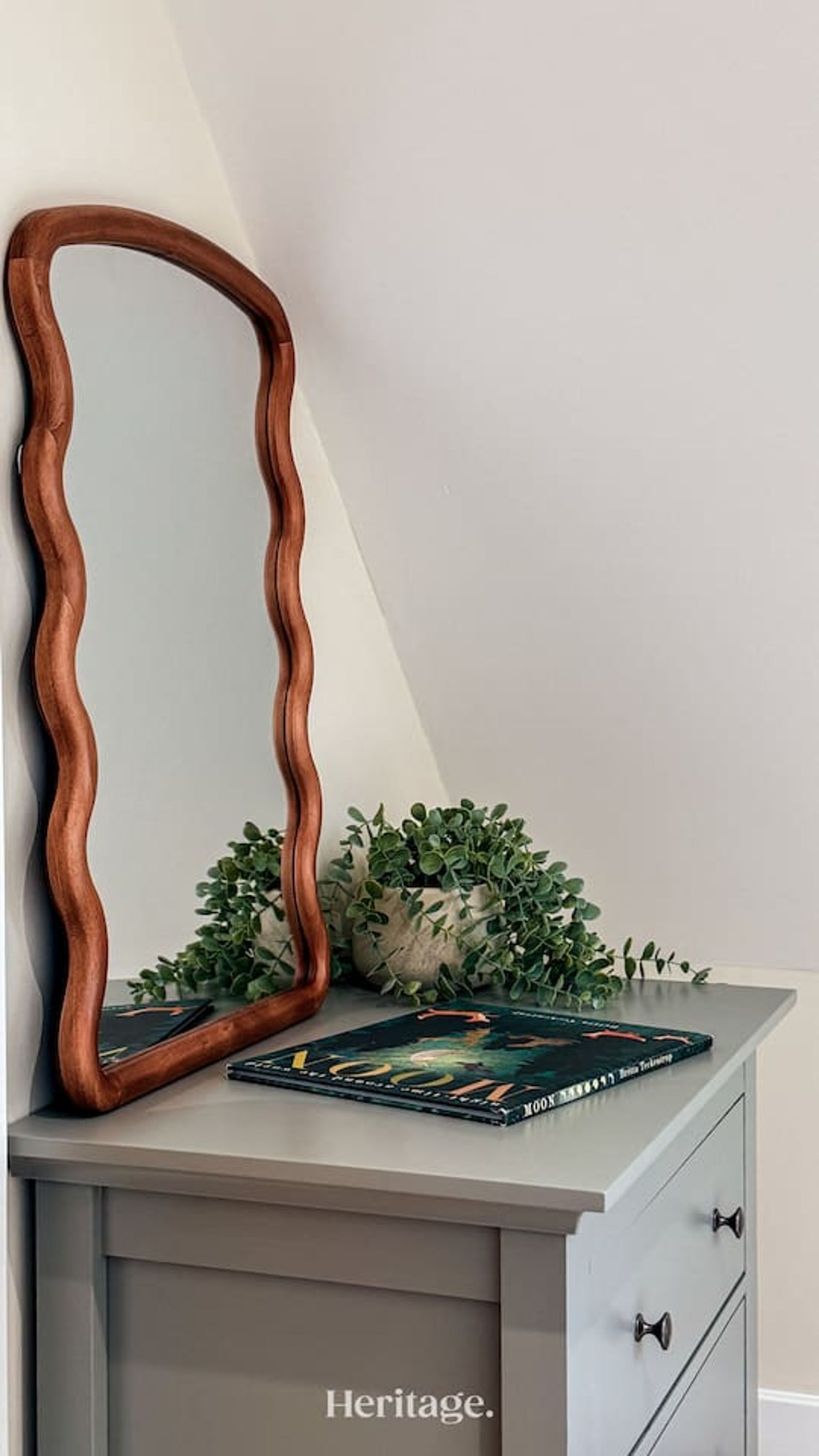 Un tocador con espejo de madera ondulado, un libro titulado 'Room' y una pequeña planta en maceta.