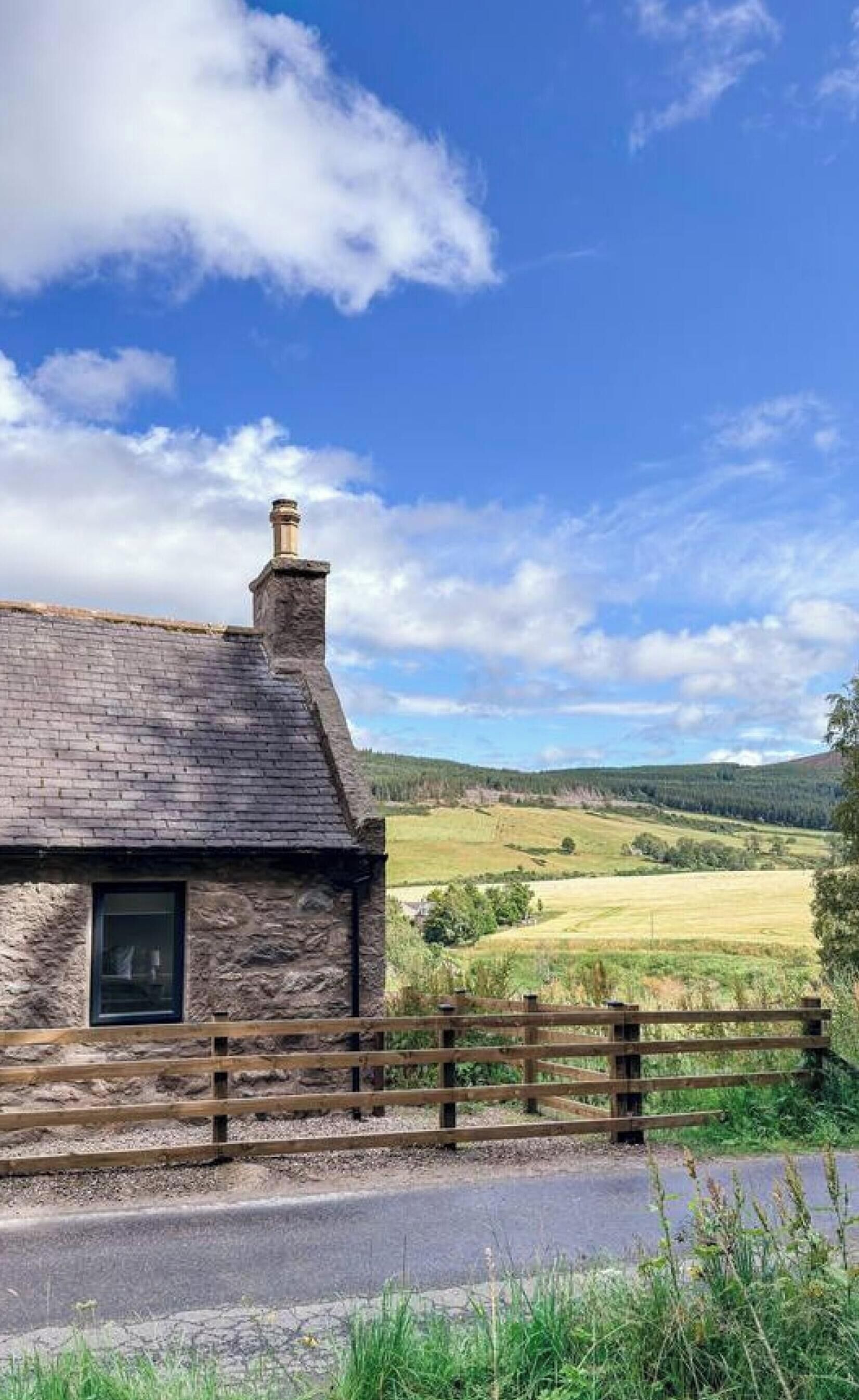 A stone cottage with a wooden fence, a chimney, and a view of a green landscape.