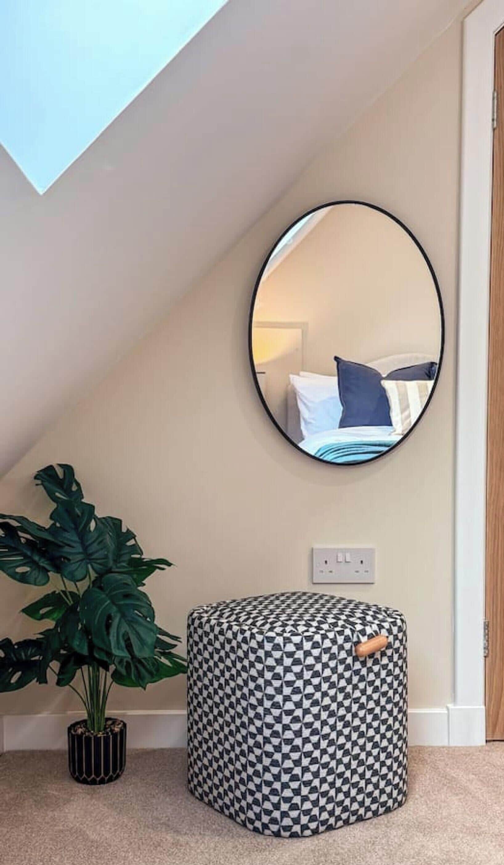 A room with a round mirror on the wall, a potted plant, and a checkered ottoman.