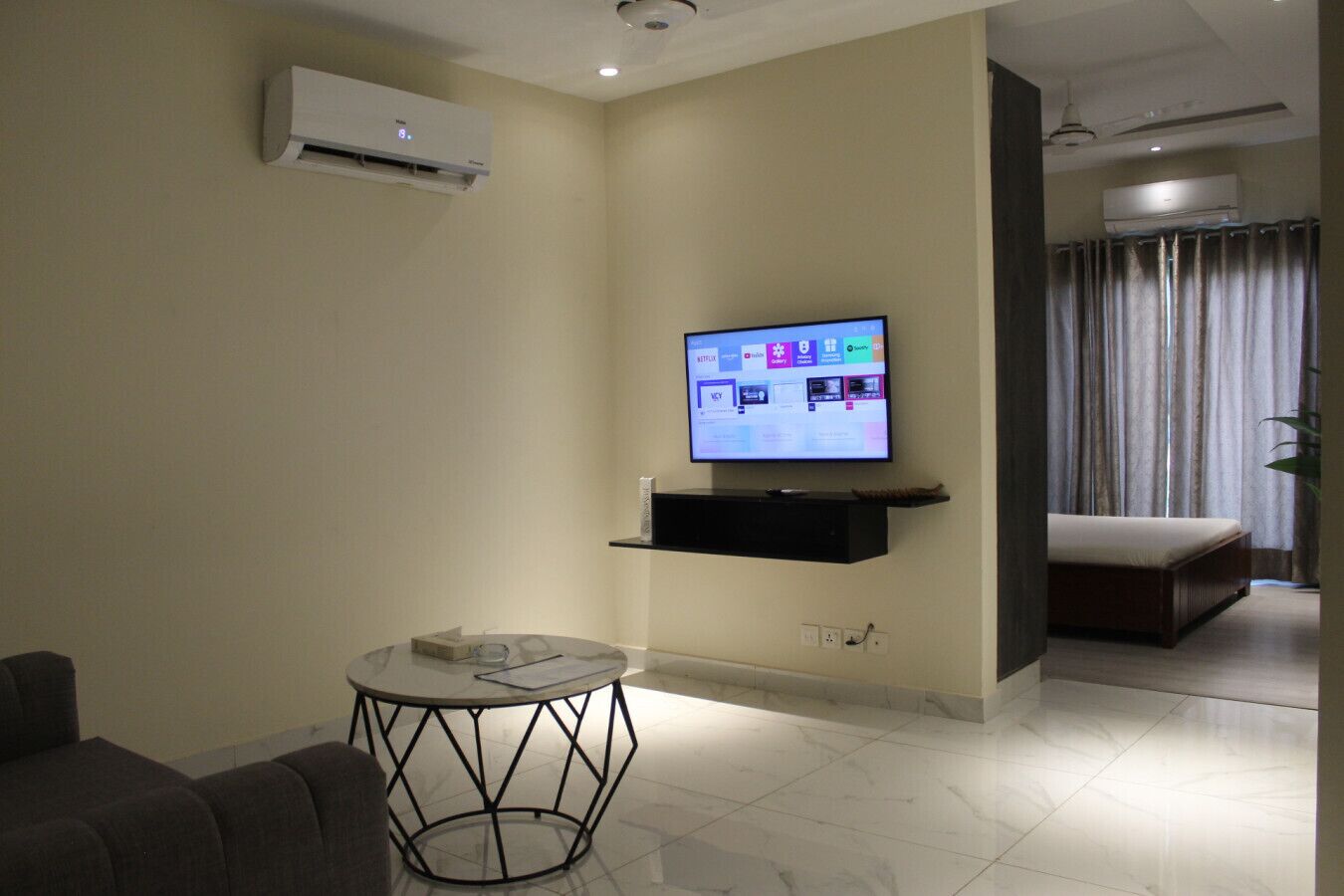 Una sala moderna con un televisor de pantalla plana montado en la pared, una mesa redonda, un sofá y una cama en la habitación contigua.