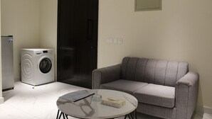A room with a washing machine, a dark door, a grey sofa, and a small round table with a box and a remote control. - Studio Apt | Roof Garden | The Opus (Lahore)