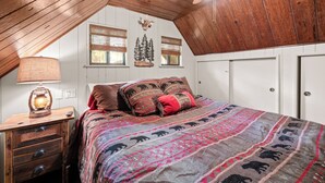 A bedroom with a sloped ceiling, a bed with patterned bedding, a lamp, and a wall-mounted clock. - Disney Imagineer-Built Cabin, Pet Friendly, Fire Pit, Walk to Town Trails (Big Bear Lake)