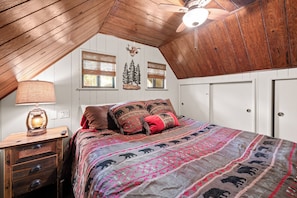 A bedroom with a sloped ceiling, a bed with patterned bedding, a lamp, and a wall-mounted clock. - Disney Imagineer-Built Cabin | Pet Friendly | Fire Pit | Walk to Town Trails (Big Bear Lake)
