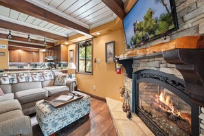 A cozy living room with a fireplace, a sofa set, and a kitchen area visible through an open door. - Disney Imagineer-Built Chalet Near the slopes | Fire Pit, Trails | Hot Tub (Big Bear Lake)
