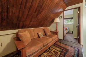 Ein gemütliches Zimmer mit Holzbalkendecke, einem gemusterten Sofa, einer Lampe und Blick auf ein weiteres Zimmer durch eine geöffnete Tür.