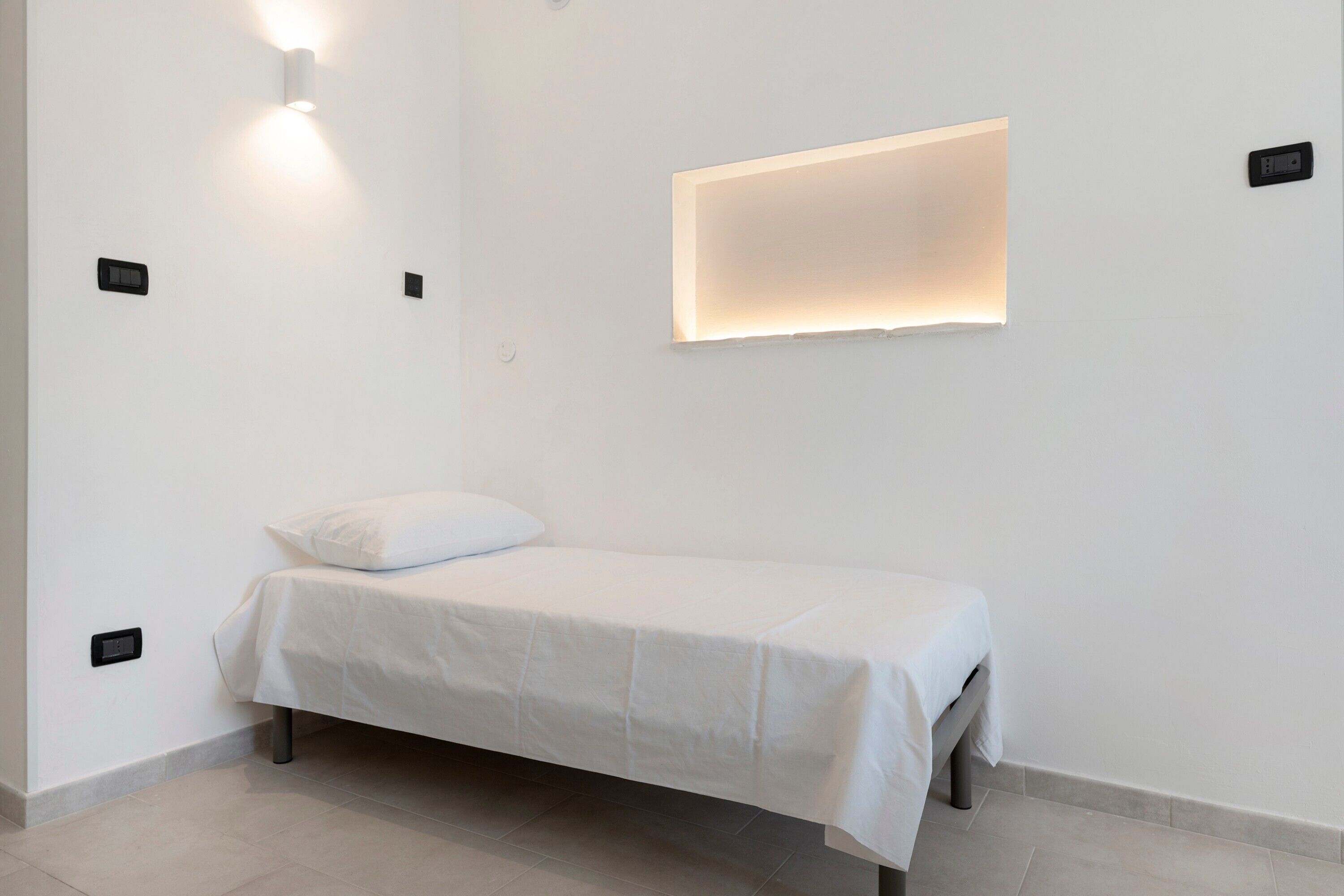 A minimalist room with a single bed, a wall-mounted light, and a recessed ceiling light.