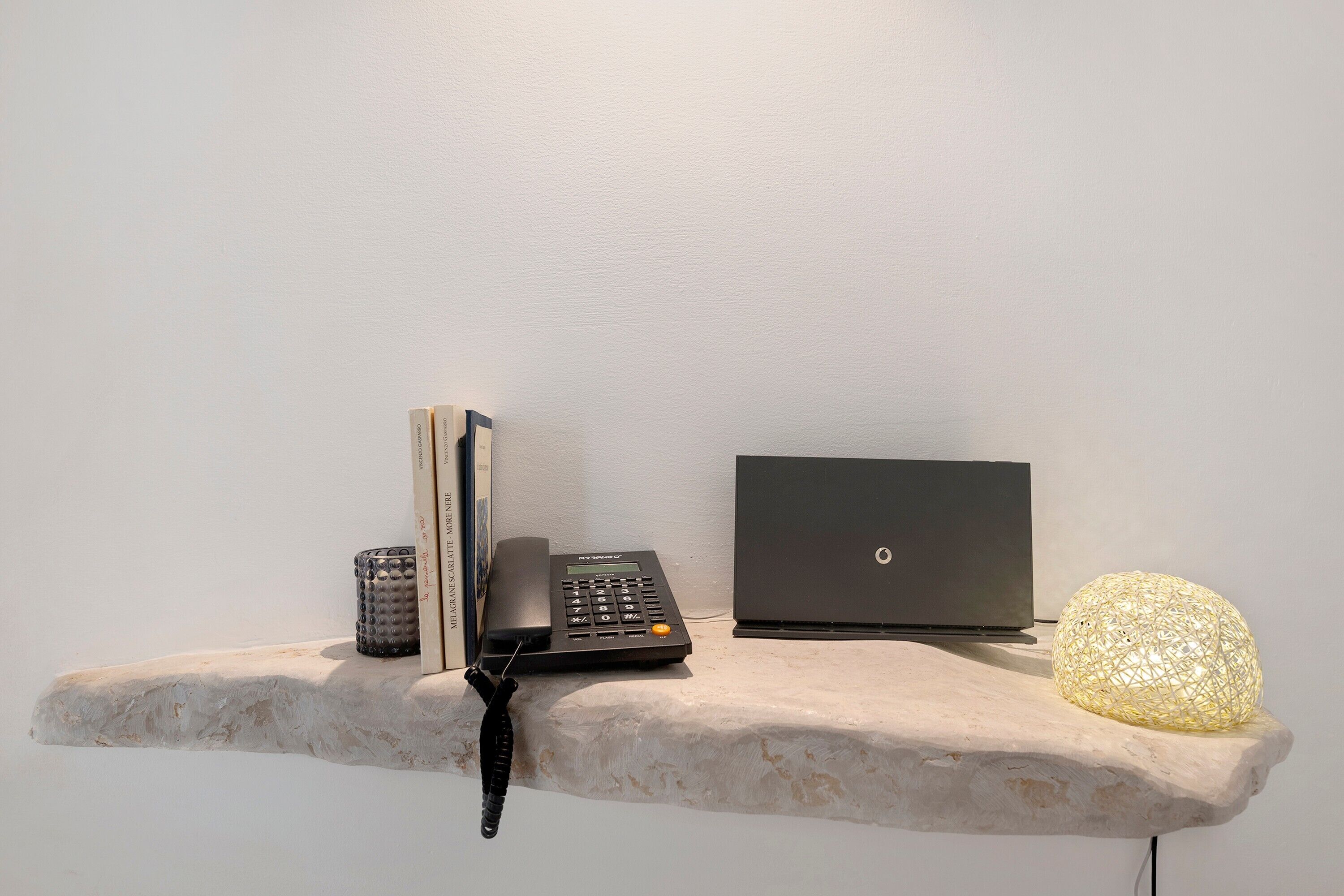A desk with a laptop, a telephone, a decorative lamp, and a book on a stone shelf.