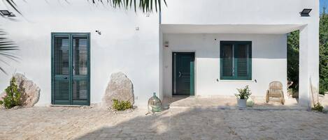 Um edifício branco com duas janelas e uma porta, uma parede de pedra e um pátio de paralelepípedos.