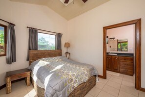 Un dormitorio con cama, mesita de noche, lámpara, ventana con cortinas y un baño con espejo y lavamanos.