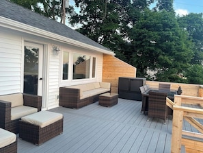 Eine überdachte Terrasse im Freien mit Korbmöbeln, eine Holzterrasse und ein Gebäude mit weißen Fenstern.