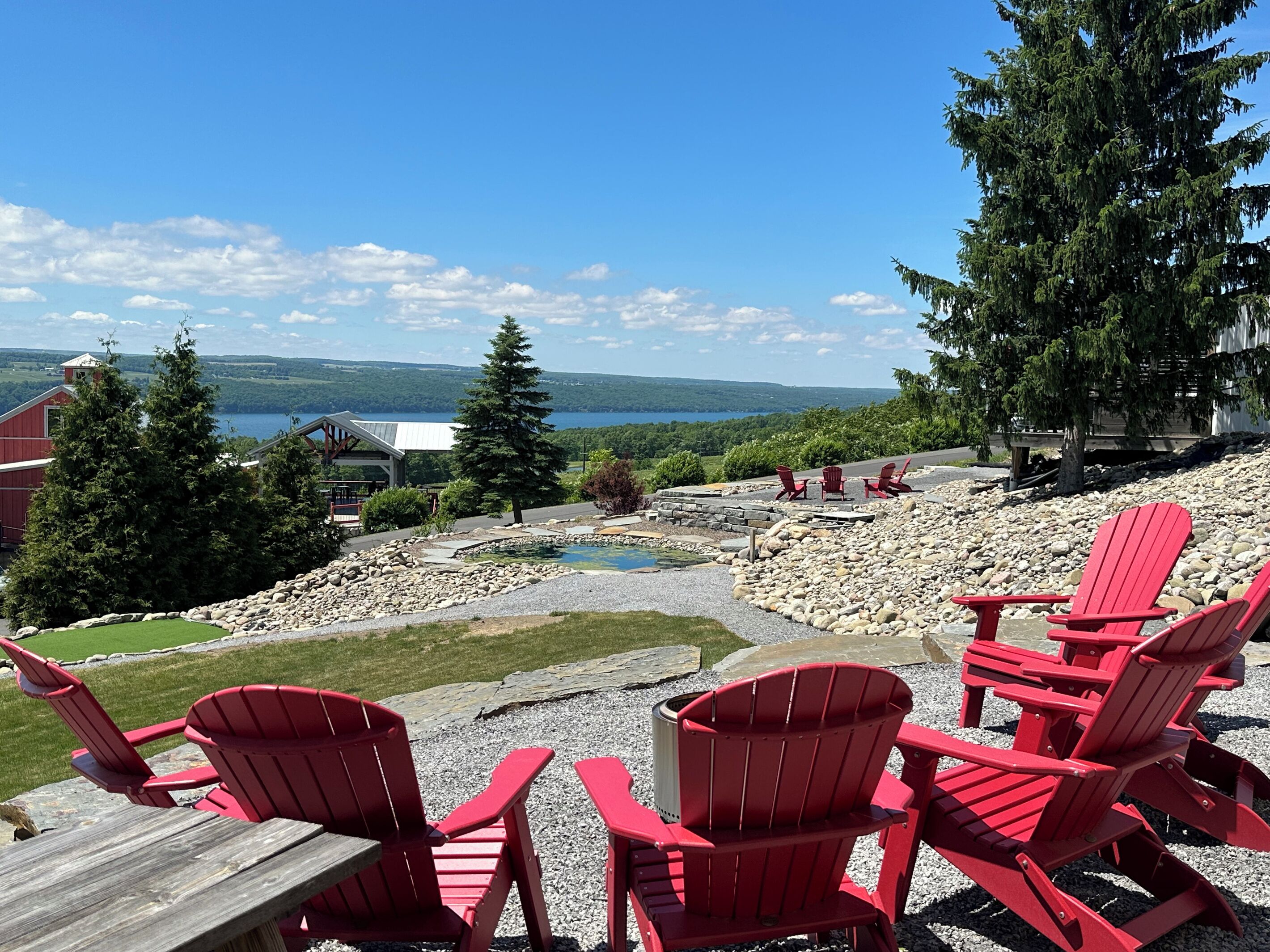 En terrass med röda Adirondack-stolar med utsikt över en sjö, en liten damm och en avlägsen vy över en röd byggnad och träd.
