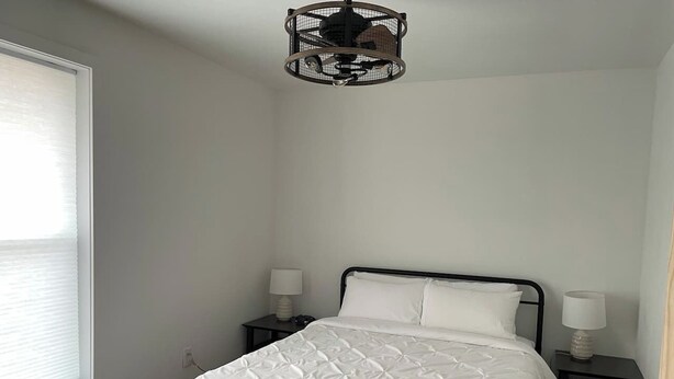 Ein Einzelbett mit Metallgestell, zwei Nachttische mit Lampen, ein Fenster mit Rollo und eine Pendelleuchte.
