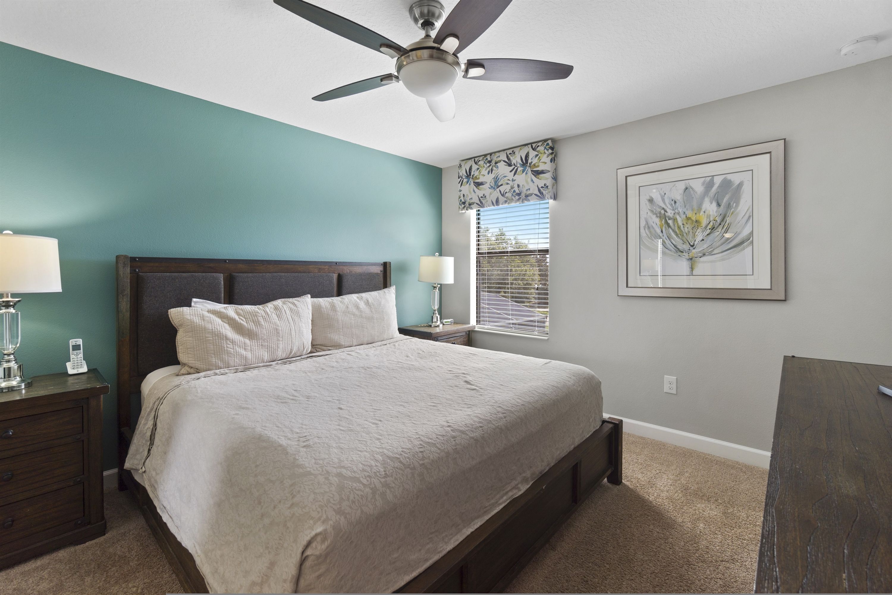 A bedroom with a bed, nightstands, a ceiling fan, a window with blinds, and a framed artwork on the wall.