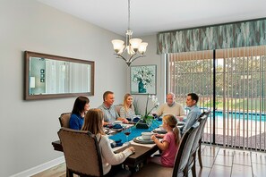 Une salle à manger familiale avec une grande table, des chaises, un lustre, un tableau et une vue sur l’extérieur.
