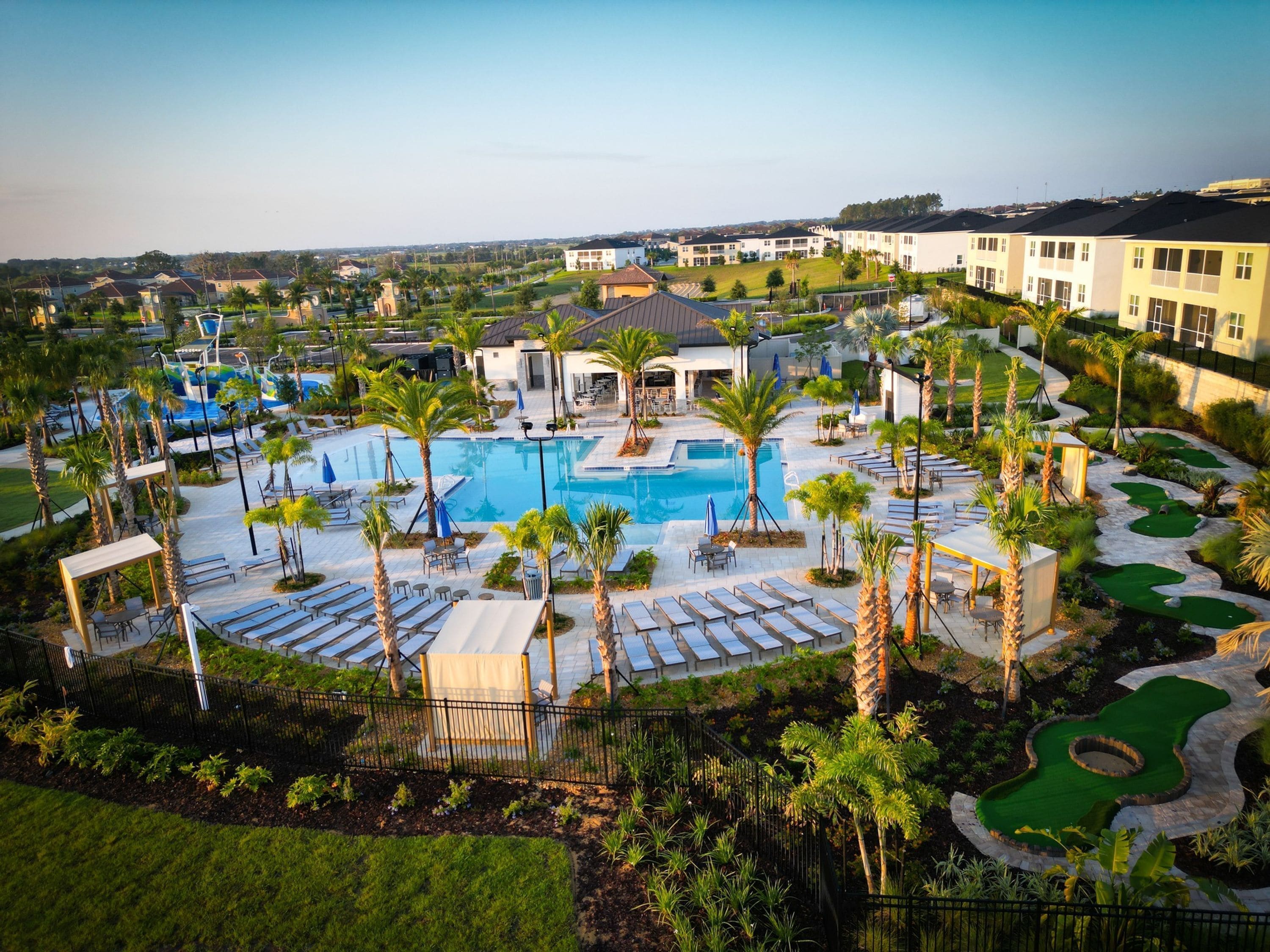 A resort with a central swimming pool, sun loungers, palm trees, and a golf course.