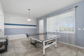 Une pièce avec une table de ping-pong, un luminaire au plafond, une fenêtre avec des stores et une porte.