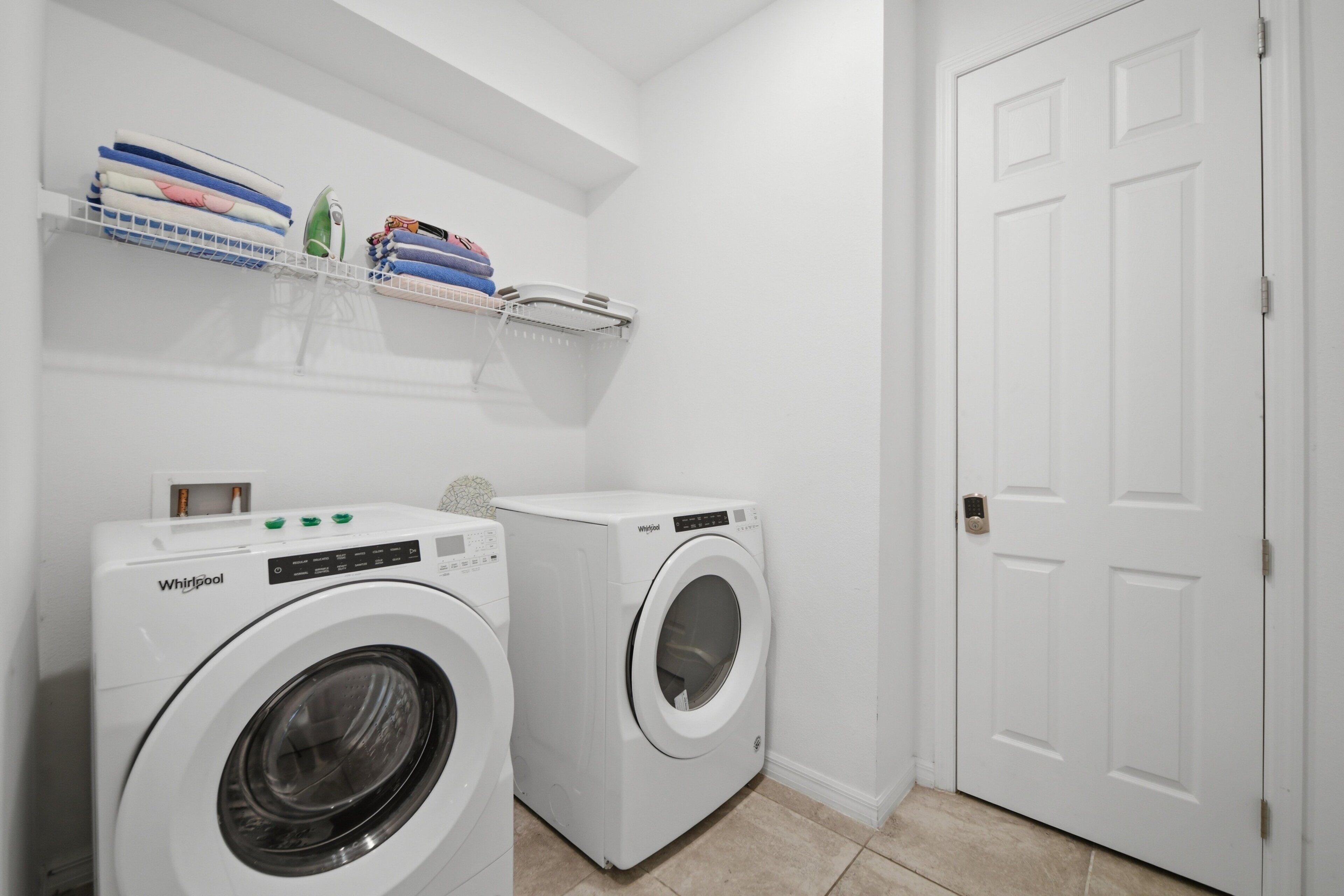 A laundry room with two Whirlpool washing machines, a white door, and a shelf with folded towels and laundry supplies.