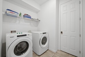 Ein Waschraum mit zwei Whirlpool Waschmaschinen, einer weißen Tür und einem Regal mit gefalteten Handtüchern und Waschutensilien.