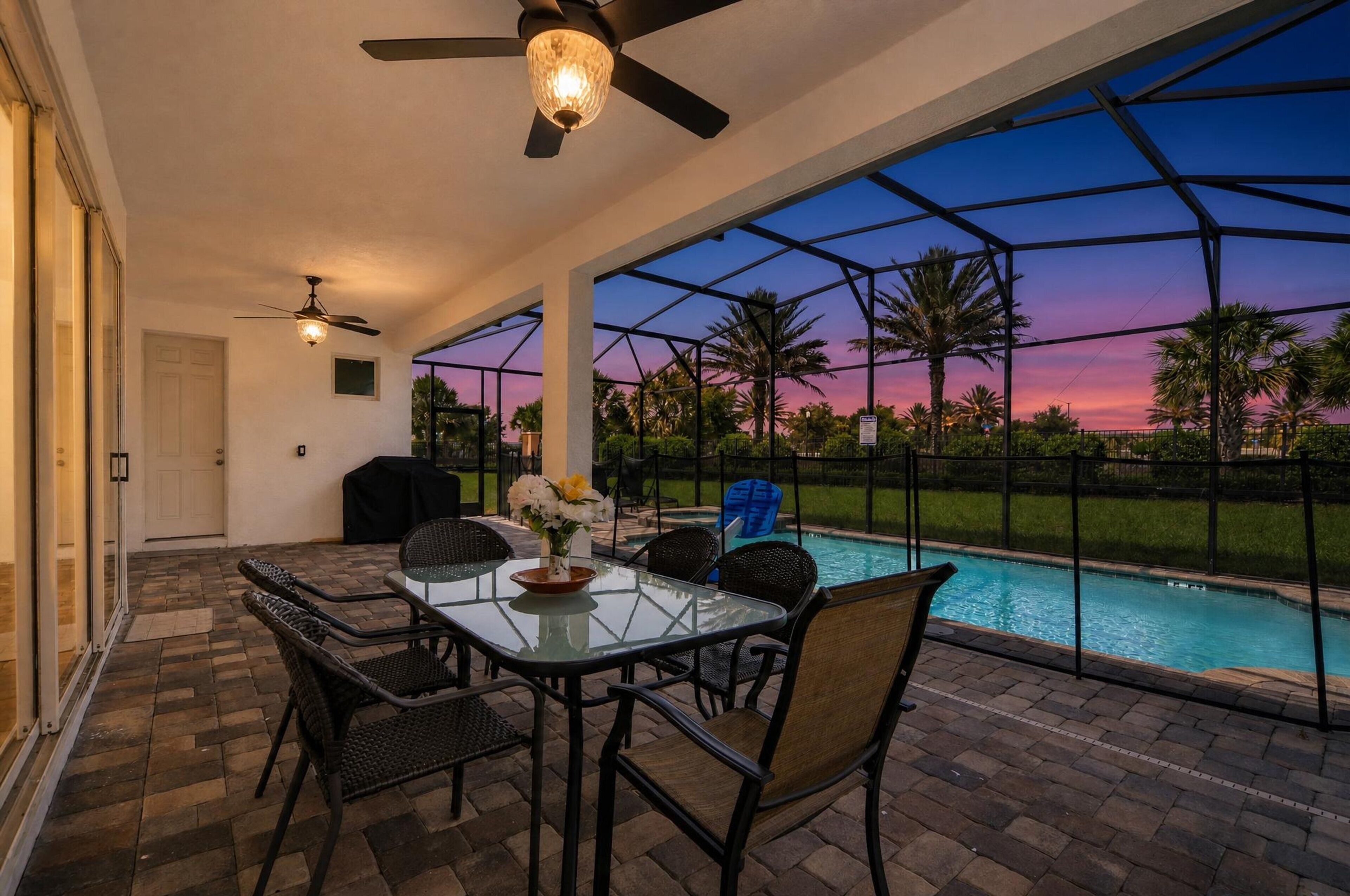 Una veranda esterna con tavolo e sedie in vetro, una ventola a soffitto e vista su una piscina al tramonto.