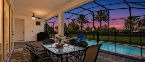 Una veranda esterna con tavolo e sedie in vetro, una ventola a soffitto e vista su una piscina al tramonto.