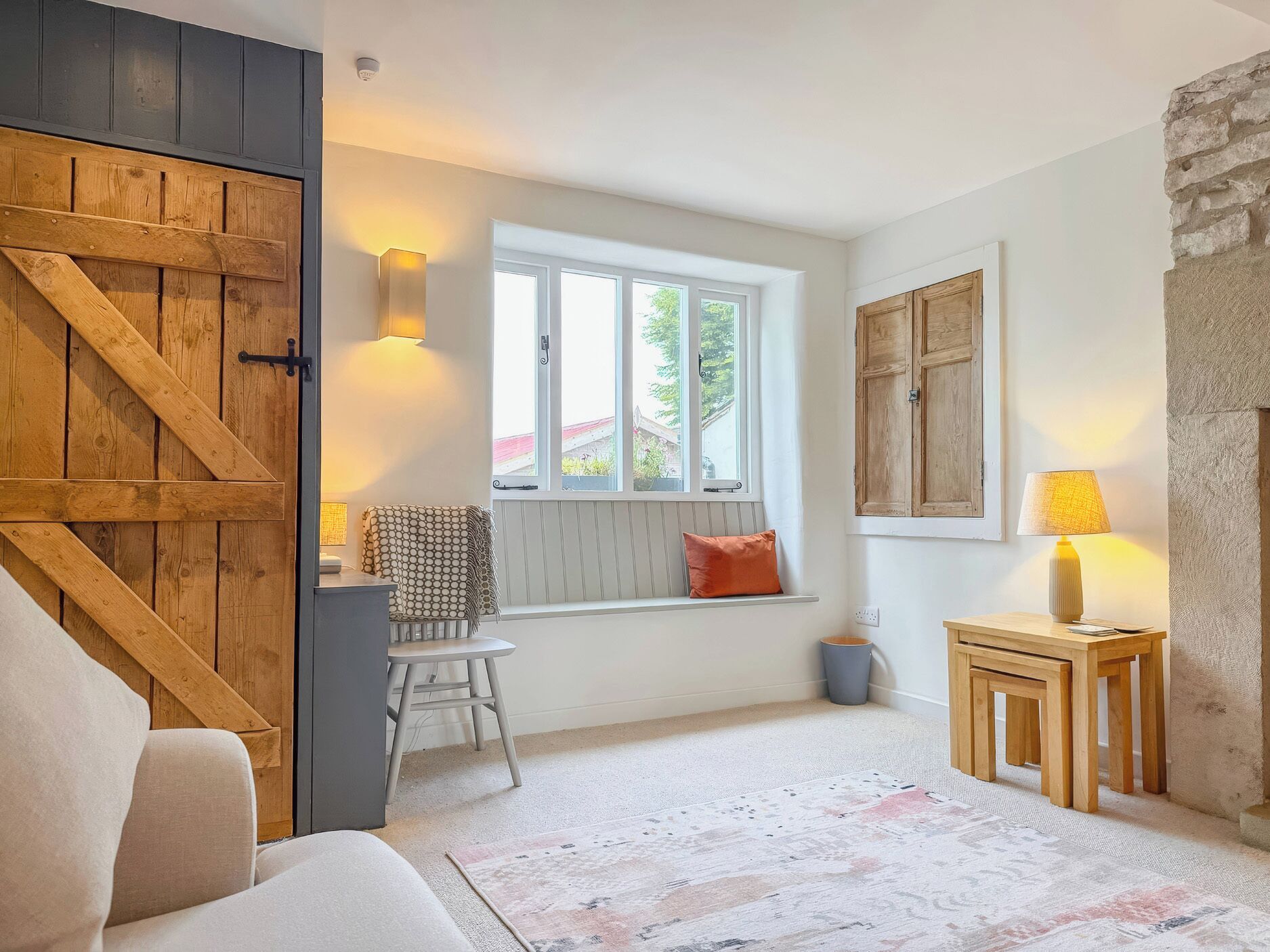 A cozy room with a large wooden door, a window with a view, a small table with a lamp, and a bench with cushions.
