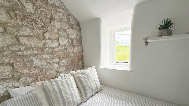 A bedroom with a stone wall, a window, a bed with striped bedding, and a small shelf with a plant.