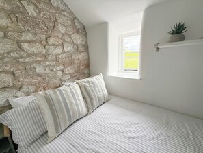 Un dormitorio con pared de piedra, una ventana, una cama con ropa de cama a rayas y un pequeño estante con una planta.