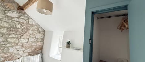 A bedroom with a stone wall, a bed with striped bedding, a hanging light fixture, and a door leading to a closet with hanging clothes.