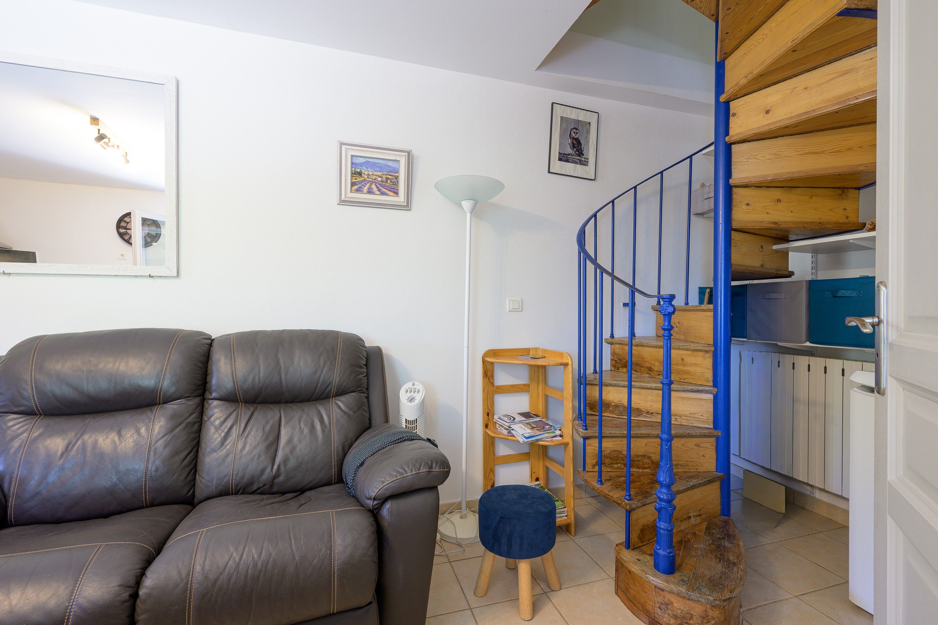 A living room with a leather sofa, a lamp, and a staircase with blue railings leading to a kitchen area.