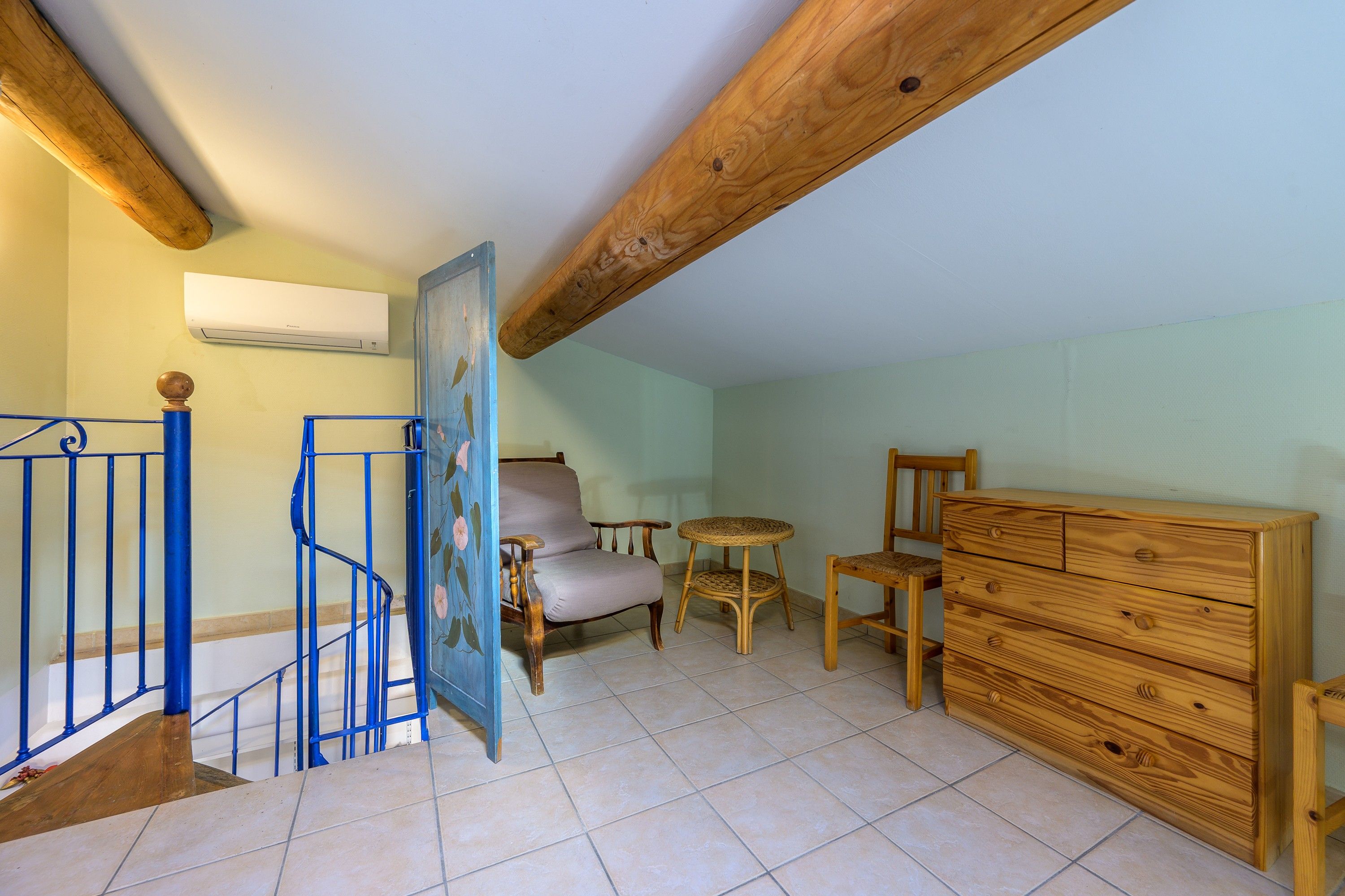 A room with a wooden staircase, a chair, a small round table, a wooden dresser, and an air conditioning unit on the wall.