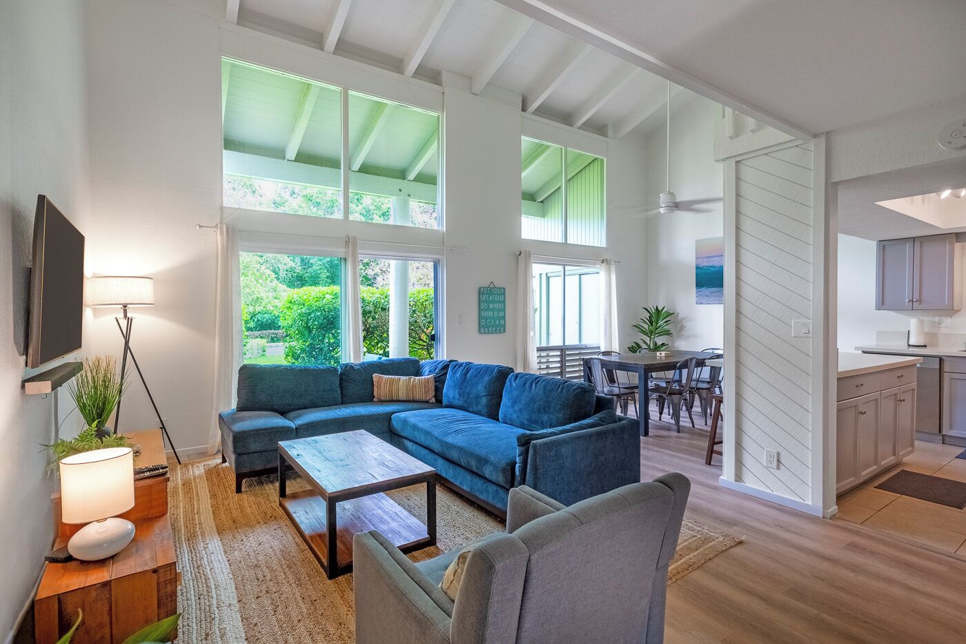 A spacious living area with a blue sofa, a wooden coffee table, and a dining table with chairs. There is a kitchen area with cabinets and a countertop visible.