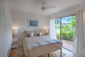 Ein Schlafzimmer mit einem Bett, Nachttischlampen, einem Deckenventilator, einem Fenster mit Vorhängen und Blick auf Grünflächen.