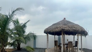 A thatched gazebo with seating, surrounded by palm trees and a pebble ground. - Beachside Studio with a Curated Library (P32W+99C Prampram)